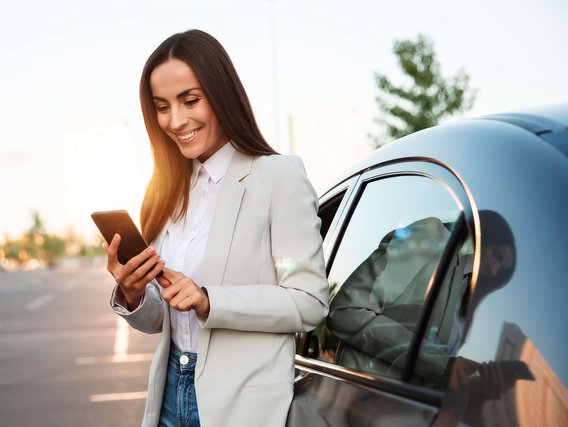 Frau steht mit dem Smartphone in der Hand neben einem Auto.