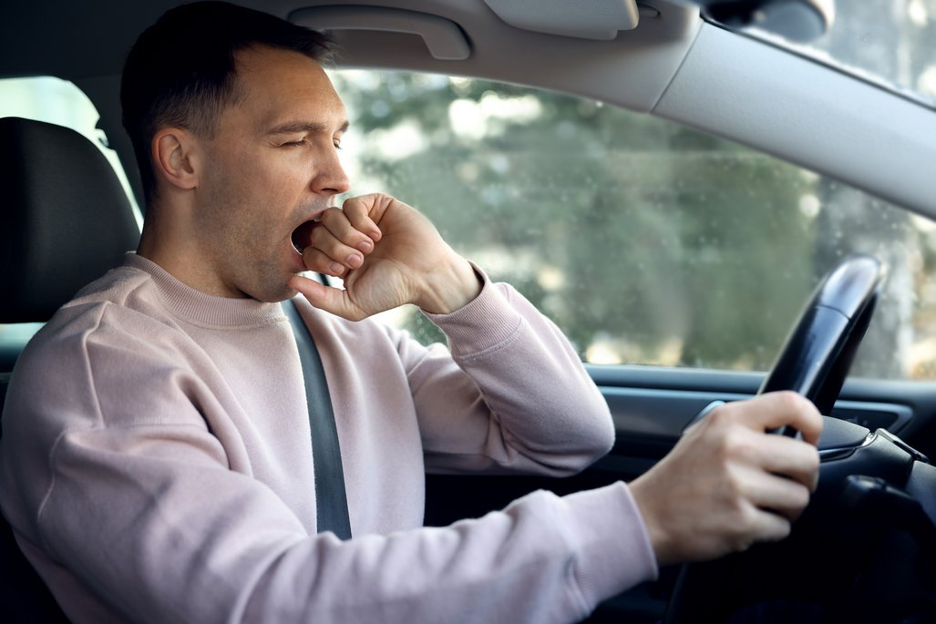 A person is seated at the wheel of a car, holding the steering wheel with one hand while suppressing a yawn with the other. The person is wearing a light-colored sweater and is buckled in. Blurred trees are visible through the window.