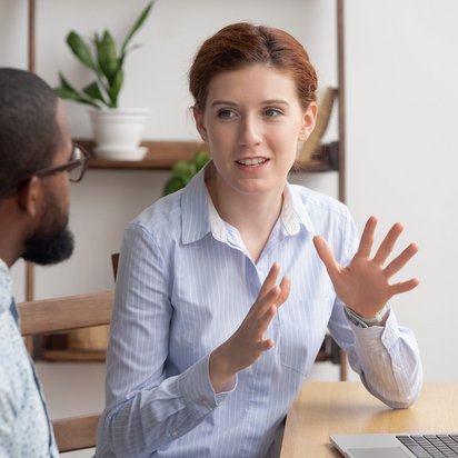 Zwei Personen in einem Gespräch am Tisch, eine Person erklärt etwas mit Gesten, während sie auf ein Laptop schaut