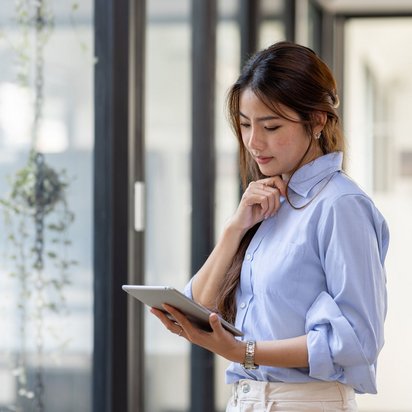 Junge Frau in hellblauer Bluse steht in einem modernen Bürogebäude und nutzt ein Tablet