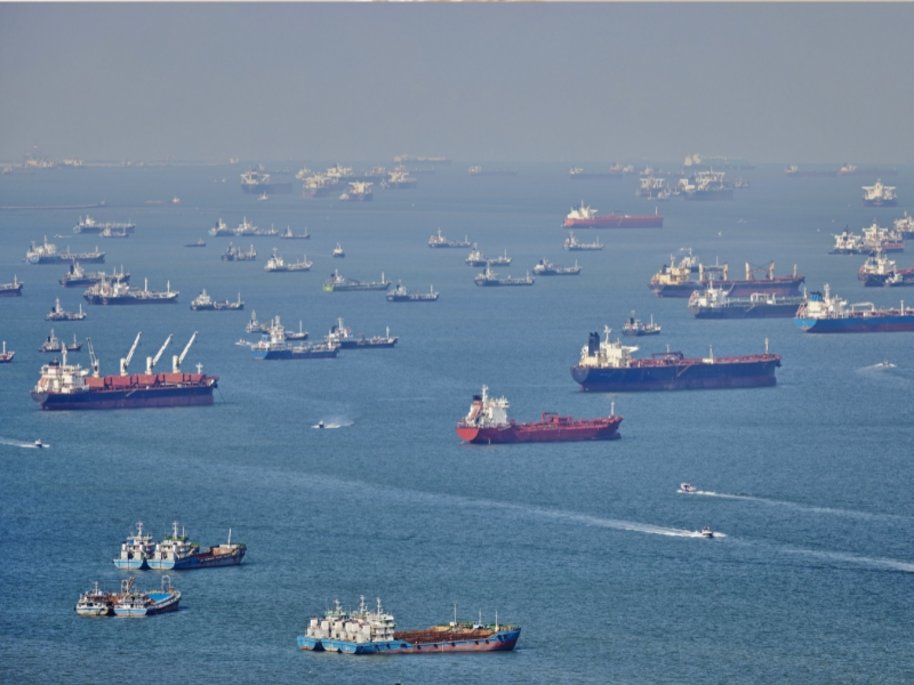 Eine Luftaufnahme eines belebten Meeresgebiets mit Dutzenden von Frachtschiffen und Tankern, die auf blauem Wasser ankern oder fahren. Die verschiedenfarbigen Schiffe sind über die gesamte Wasserfläche verteilt, einige mit roten Rümpfen sind besonders gut zu erkennen.