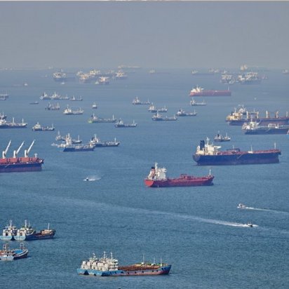 Eine Luftaufnahme eines belebten Meeresgebiets mit Dutzenden von Frachtschiffen und Tankern, die auf blauem Wasser ankern oder fahren. Die verschiedenfarbigen Schiffe sind über die gesamte Wasserfläche verteilt, einige mit roten Rümpfen sind besonders gut zu erkennen.