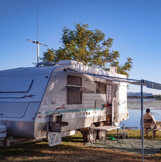 Zwei Personen sitzen an einem See bei blauem Himmel. Ein Wohnwagen steht auch auf der Wiese.