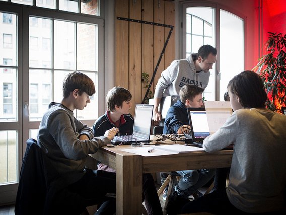 Ein Lehrer in "HACKER SCHOOL" Pullover steht hinter vier Jugendlichen, die an Laptops an einem Holztisch arbeiten.