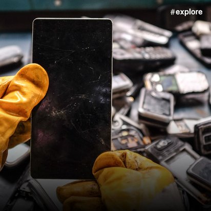 Eine Person mit gelben Arbeitshandschuhen hält ein schwarzes Smartphone vor einer ungeordneten Menge alter Mobiltelefone. Im Hintergrund sind verschiedene ausrangierte Handymodelle zu erkennen.