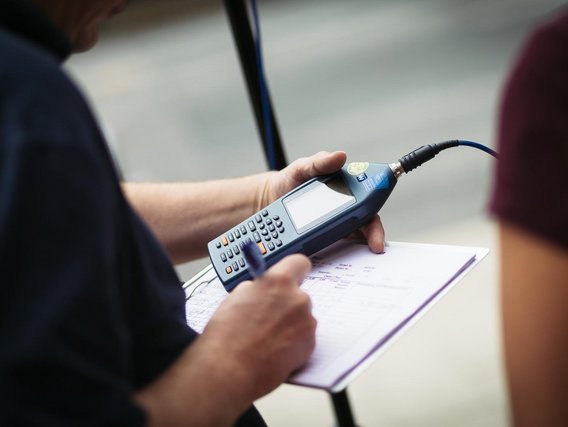 Detailaufnahme einer Hand, die ein Schallpegelmessgerät über einem Formular hält, verbunden mit einem Audiokabel.