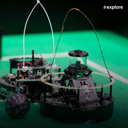 Eine Nahaufnahme eines kleinen runden Roboters mit Antennen auf einem hellgrünen Untergrund. Der Roboter hat Sensoren und Räder und ist für einen Robotik-Wettbewerb konzipiert.