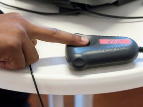 Eine Hand berührt einen grauen Fingerabdruckscanner mit der Aufschrift "NOP007" auf einer roten Anzeige.