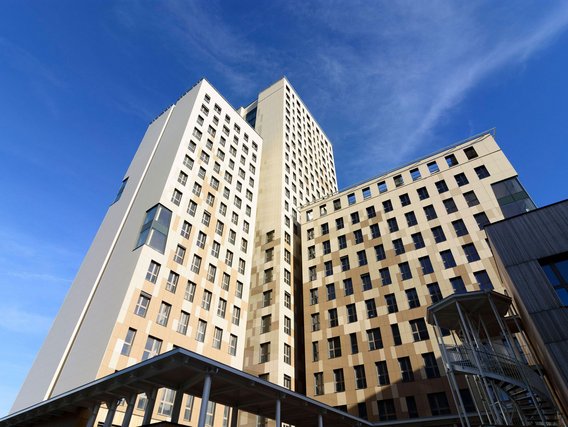 Moderne Hochhäuser in hellen Beige- und Weißtönen mit regelmäßigen Fenstern ragen gegen einen strahlend blauen Himmel empor, im Vordergrund ein Glasüberdach.