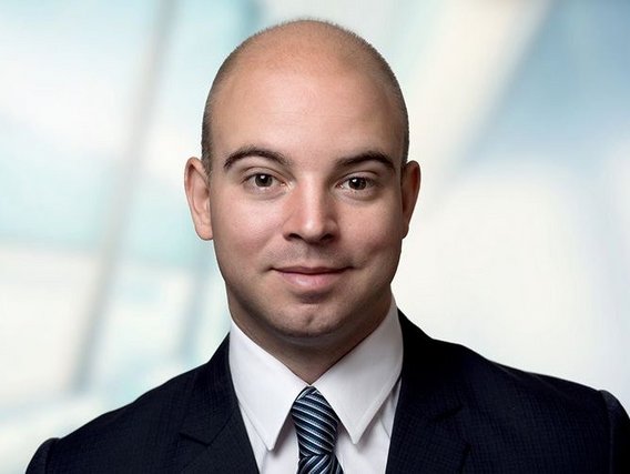 Dennis Dal Mas mit weißem Hemd, blauer Krawatte und schwarzem Anzug vor einem weißen und hellen Hintergrund.