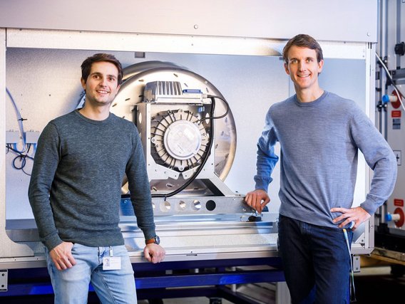 Marius Ciucas (links) und Timo Reisner in grauen Pullovern stehen vor einer geöffneten Technikeinheit mit sichtbarem Ventilator und elektronischen Komponenten.