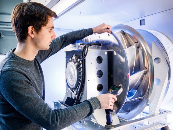 Ein Techniker mit grauem Pullover prüft mit einem Messgerät eine runde Lüftereinheit mit metallischen Bauteilen.