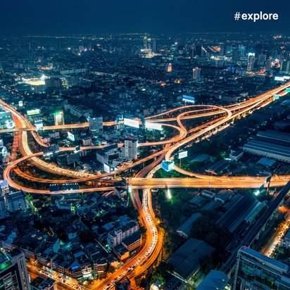 Eine lebendige Stadtlandschaft bei Nacht, die komplexe Autobahnkreuzungen und beleuchtete Gebäude zeigt. Die Straßen sind von Lichtern der Fahrzeuge erhellt, und der Hashtag "#explore" ist oben rechts im Bild zu sehen.
