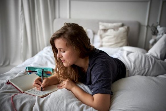 Eine junge Frau mit langen braunen Haaren liegt in bequemer Kleidung auf einem Bett mit weißen Laken und schreibt in ihr Notizbuch.