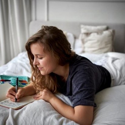 Eine junge Frau mit langen braunen Haaren liegt in bequemer Kleidung auf einem Bett mit weißen Laken und schreibt in ihr Notizbuch.