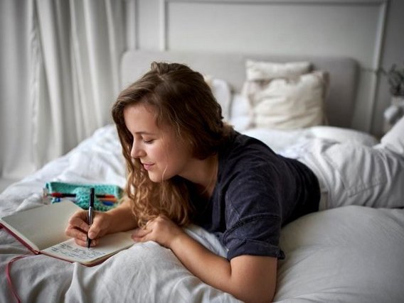 Eine junge Frau mit langen braunen Haaren liegt in bequemer Kleidung auf einem Bett mit weißen Laken und schreibt in ihr Notizbuch.