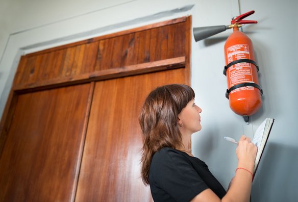 Frau überprüft Feuerlöscher an der Wand und notiert Daten in ein Klemmbrett
