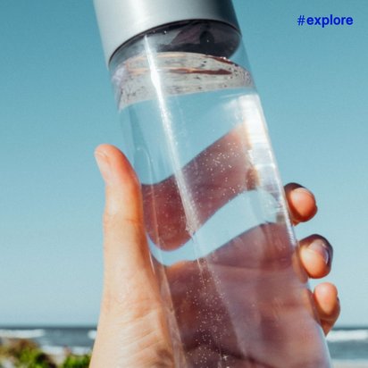 Eine Hand hält eine halb gefüllte Glaswasserflasche gegen blauen Himmel mit unscharfer Küstenlandschaft im Hintergrund.
