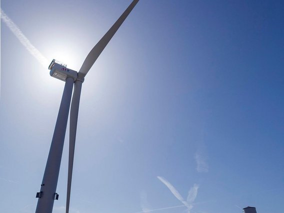 A large offshore wind turbine with white tower and nacelle stands on a bright yellow floating platform foundation at a port facility. The turbine is positioned on land with installation equipment visible nearby under clear blue skies.