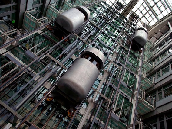 Modernes Glasaufzugsystem im Atrium eines zeitgenössischen Gebäudes, das mehrere zylindrische Aufzugskabinen zeigt, die an freiliegenden Metallschienen vor einer Glas- und Stahlarchitektur montiert sind.