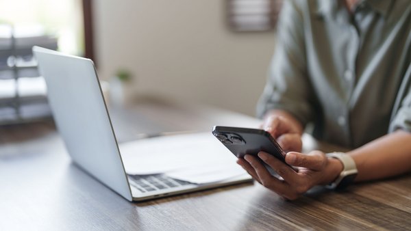 Person hält ein Smartphone in der Hand, vor ihr steht ein aufgeklappter Laptop auf einem Schreibtisch.