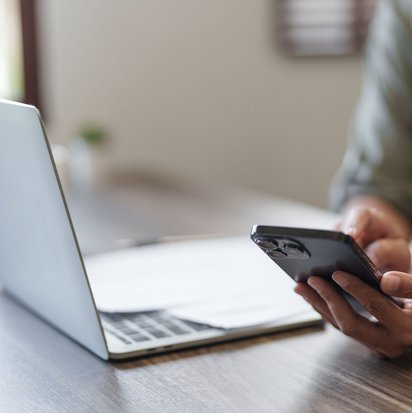 Person hält ein Smartphone in der Hand, vor ihr steht ein aufgeklappter Laptop auf einem Schreibtisch.