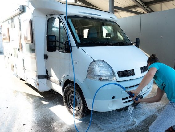 Eine Person in türkisfarbenem Shirt wäscht ein weißes Wohnmobil mit blauem Wasserschlauch in einer überdachten Waschstation.