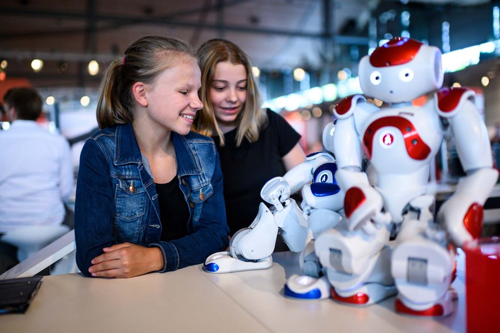 Zwei junge Mädchen interagieren mit weißen und roten Robotern an einem Messestand. Die Mädchen schauen begeistert auf die Roboter, die auf einem Tisch präsentiert werden.
