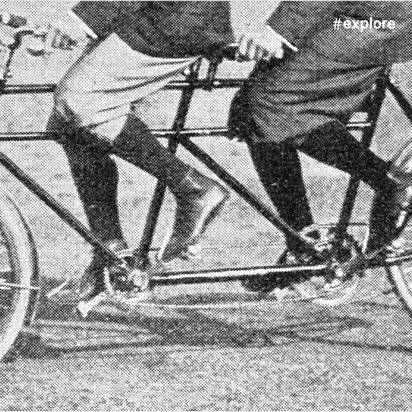 Schwarzweißfoto eines historischen Tandemfahrrads mit zwei Fahrern. Die komplexe Konstruktion zeigt die technische Innovation der Frühzeit des Radsports.