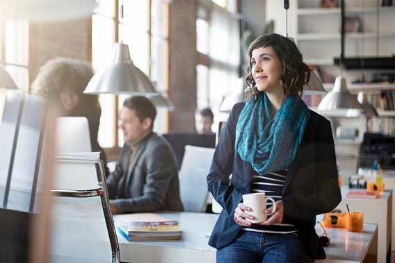 Eine Frau lächelt und hält eine Tasse in den Händen, im Hintergrund arbeiten weitere Personen im Büro.