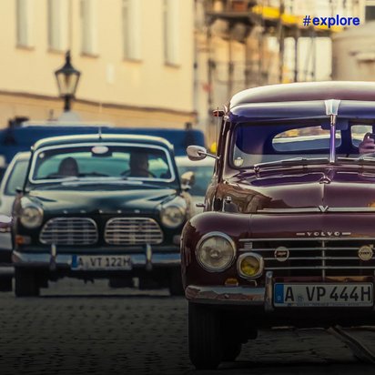 Ein bordeauxroter klassischer Volvo-Oldtimer parkt auf Kopfsteinpflaster in einer europäischen Altstadt. Im Hintergrund sind weitere Autos und historische Gebäude mit Straßenlaternen zu sehen.