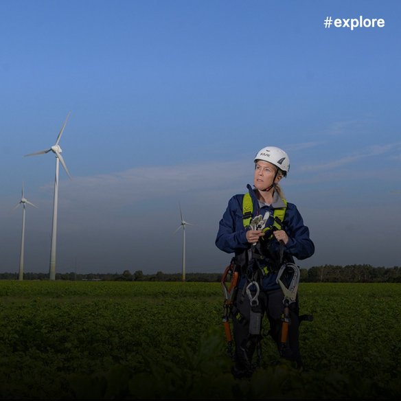 Eine Technikerin in Sicherheitsausrüstung steht vor einem Windpark. Sie hält Werkzeuge und inspiziert die Turbinen unter klarem Himmel.