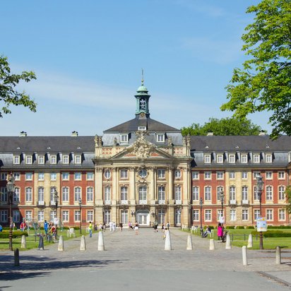 Schloss Münster mit Vorplatz und Personen bei sonnigem Wetter, eingerahmt von Bäumen