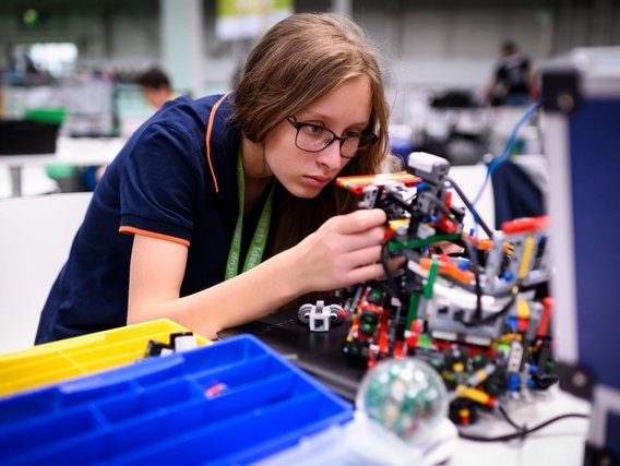 Ein Mädchen mit Brille und dunkelblaues Shirt mit grünem Lanyard arbeitet hochkonzentriert an einem komplexen LEGO-Technic-Roboter. Neben ihr sind farbige Sortierkästen für die Bauteile zu sehen.