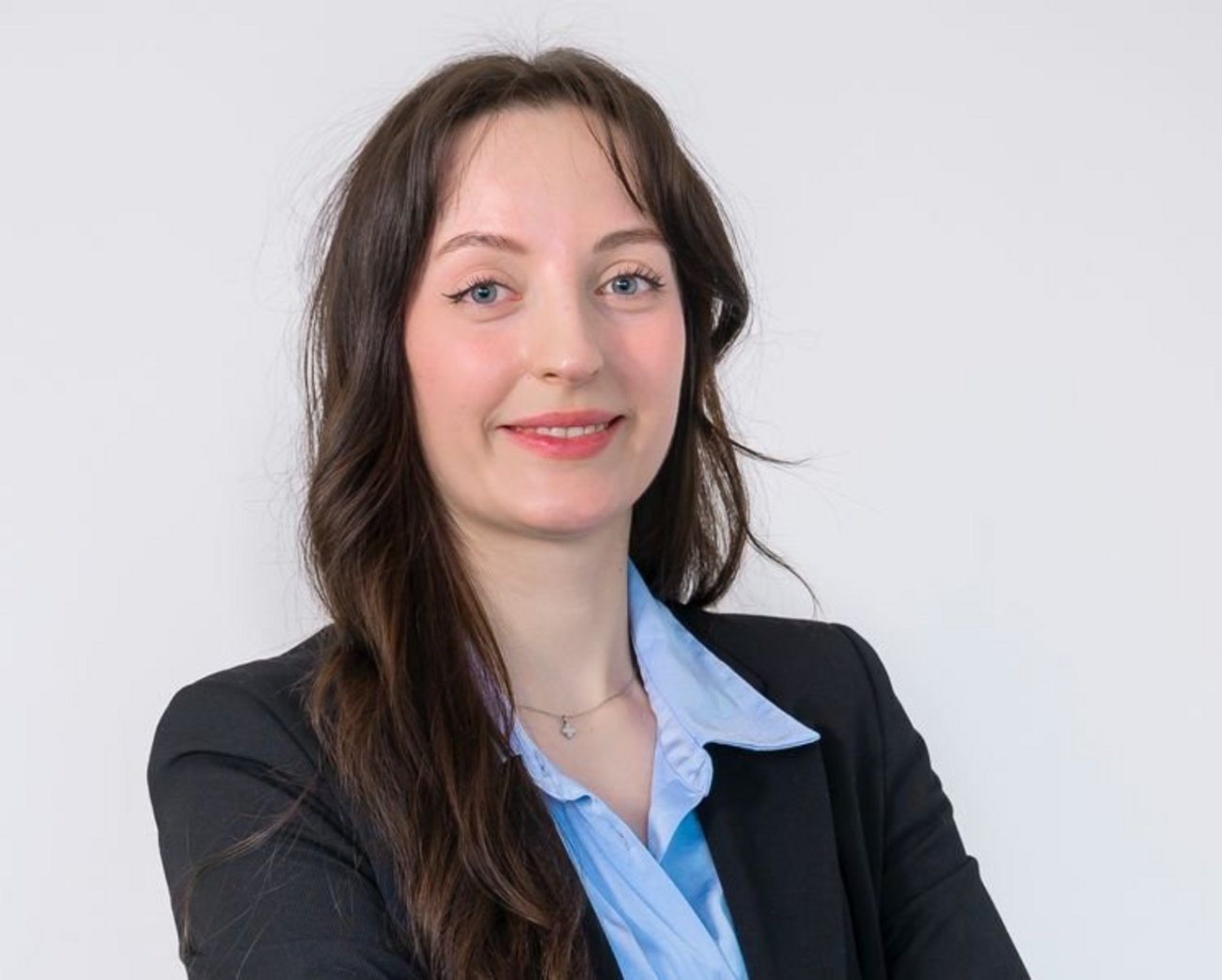 Person standing against a light background wearing a light shirt under a dark blazer, with arms loosely crossed in front.