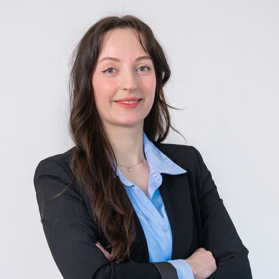 Person standing against a light background wearing a light shirt under a dark blazer, with arms loosely crossed in front.