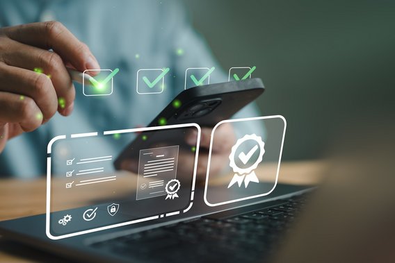 Hand mit Smartphone vor Laptop, digitale Prüf- und Zertifizierungssymbole