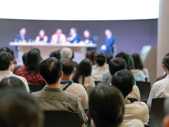 Das Bild zeigt die Rückansicht eines Publikums in einem Konferenzraum mit Speaker auf der Bühne
