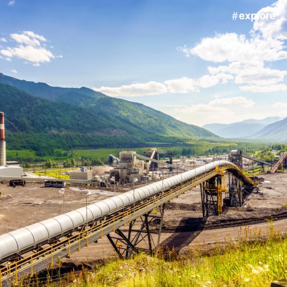 Industrieanlage in einem breiten Tal mit bewaldeten Bergen im Hintergrund. Eine lange, über Gestellen geführte Rohrleitung oder Förderstrecke verläuft diagonal durch das Gelände zu mehreren Industriegebäuden und technischen Anlagen. Der Himmel ist blau mit vereinzelten Wolken.
