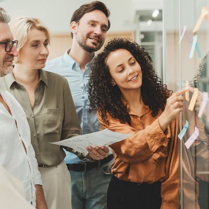 Frau befestigt farbige Zettel an einer Glaswand, umgeben von einer Gruppe lächelnder Kolleg:innen