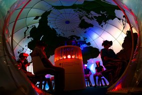 Eine immersive Erfahrungsstation mit einer Weltkarte-Projektion. Menschen mit VR-Brillen sitzen in einem kreisförmigen Raum. In der Mitte ist eine zylindrische Struktur mit roten Lichtern zu sehen. Das Logo des Bundesministeriums für wirtschaftliche Zusammenarbeit und Entwicklung ist erkennbar.