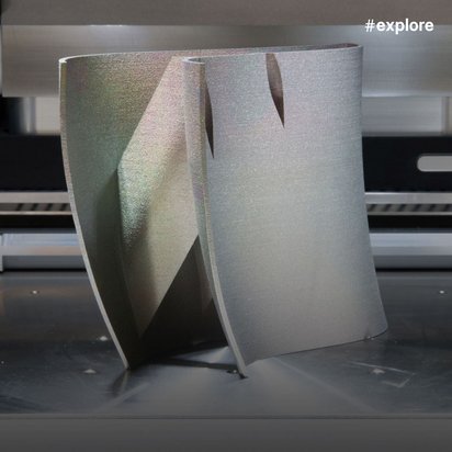 Ein 3D-Drucker druckt gerade ein graues geometrisches Objekt mit dreieckigen Formen auf einer schwarzen Druckplatte. Die Maschine zeigt präzise Mechanik mit weißen Führungsrollen und Metallschienen im Hintergrund.