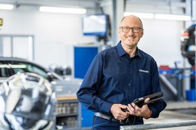 TÜV NORD Mitarbeiter mit Tablet in einer modernen Werkstatt, im Hintergrund Werkstattausrüstung und ein Auto.