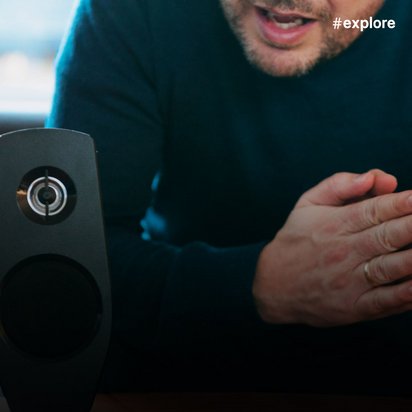 Ein Mann in dunkler Kleidung gestikuliert während eines Gesprächs oder einer Präsentation, neben ihm steht ein schwarzer Lautsprecher. Die Aufnahme zeigt ihn in einer kommunikativen Situation, möglicherweise bei einer Audio- oder Technikdemonstration.