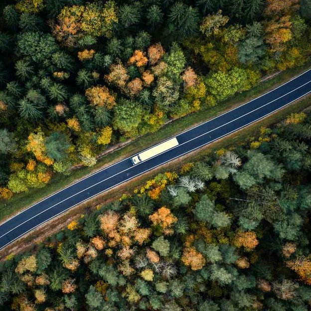 Luftaufnahme eines Lastwagens, der auf einer kurvenreichen Straße durch einen herbstlich gefärbten Wald fährt.
