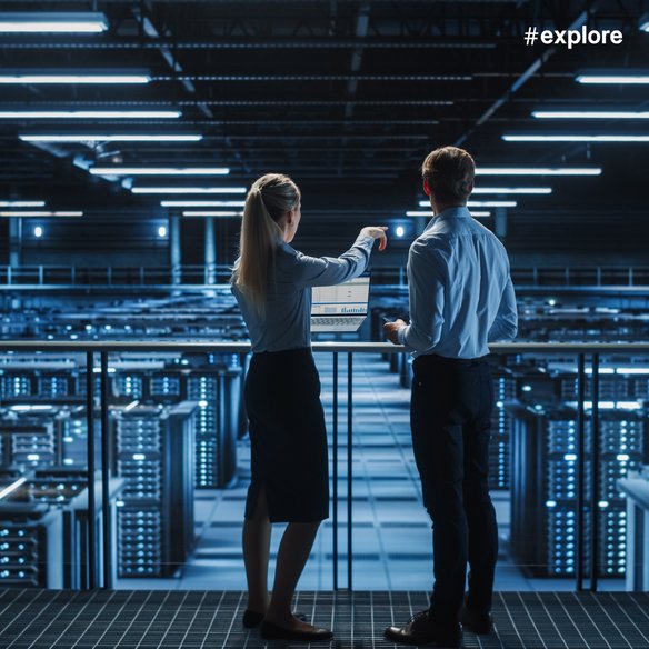 Zwei Personen stehen in einem modernen Rechenzentrum und betrachten einen Laptop mit Datenanalysen. Die Umgebung ist von blauen Lichtern und zahlreichen Servern geprägt, die eine technologische und futuristische Atmosphäre schaffen.