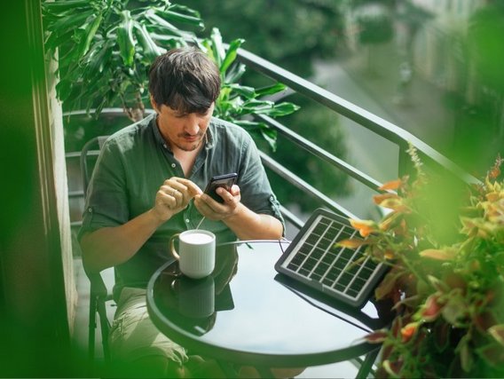 Ein Mann in einem grünen Hemd sitzt auf einem Balkon mit Pflanzen, benutzt sein Smartphone und hat einen Laptop und eine Tasse auf dem Tisch vor sich.