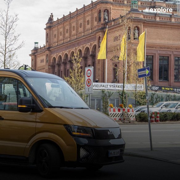 Goldener MOIA-Elektrobus vor einem imposanten historischen Backsteingebäude mit Arkaden und gelben Fahnen, umgeben von Baustellen-Absperrungen.