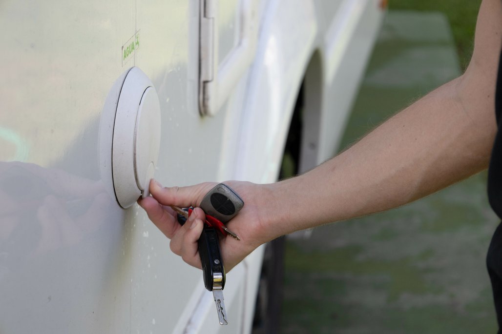 Eine Hand hält einen schwarzen Fahrzeugschlüssel vor die weiße Außentür eines Wohnmobils oder Caravans. Am Handgelenk ist eine dunkle Uhr oder ein Fitness-Tracker zu sehen.