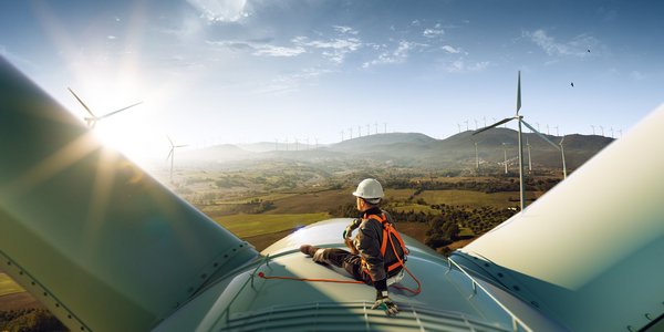 Techniker mit Sicherheitsausrüstung sitzt auf einer Windkraftanlage mit Blick auf ein Tal und weitere Windräder.